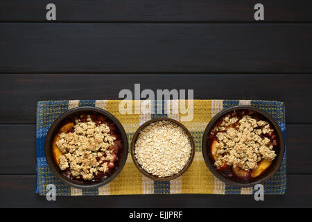Overhead shot of two rustic bowls filled with baked plum and nectarine crumble or crisp and a bowl of rolled oats Stock Photo