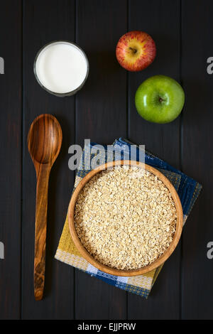 Overhead shot of raw rolled oats in wooden bowl with apples and a glass of milk, photographed on dark wood with natural light Stock Photo