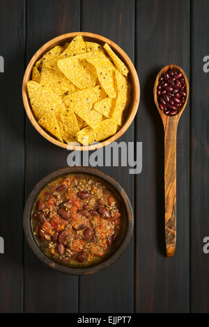 Overhead shot of chili con carne and tortilla chips in bowls with dried kidney beans on wooden spoon, photographed on dark wood Stock Photo