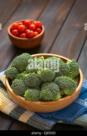 Fresh raw broccoli florets in wooden bowl with cherry tomatoes in the back, photographed on dark wood with natural light Stock Photo