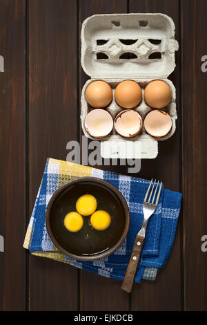 Overhead shot of three raw eggs in rustic bowl with fork on the side and egg box with eggs and eggshells above Stock Photo