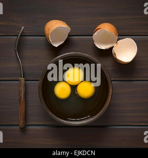 Overhead shot of three raw eggs in rustic bowl with fork on the side and broken eggshells above Stock Photo