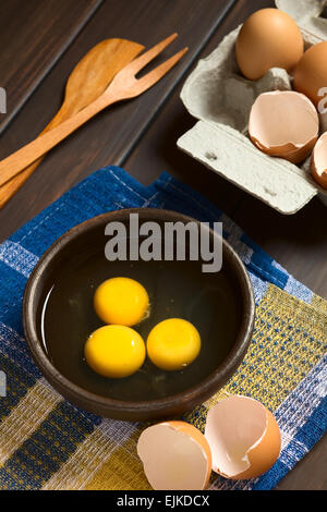 Three raw eggs in rustic bowl with egg box with eggs and eggshells in the back, photographed on dark wood with natural light Stock Photo