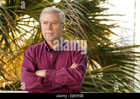 Senior man in his 60s standing with arms crossed in front of a palm Stock Photo