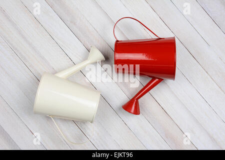 Two watering cans, one white another red laying on their side on a rustic whitewashed wood surface. Stock Photo