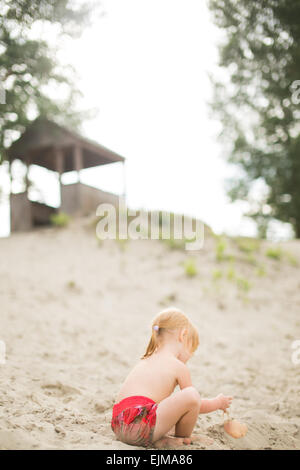 Cute toddler girl with red hair at the beach playing with sand. Child in pink shorts sitting on sand. Wooden cabin on the backgr