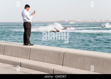 A man stands on the waterfront taking a cell phone picture a jet-ski competition in Doha, Qatar. Stock Photo
