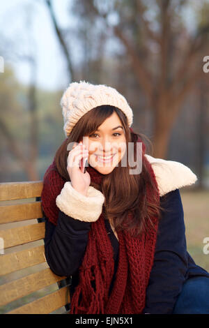 Smiling girl talking on mobile phone on bench in park in the Winter looking at camera Stock Photo