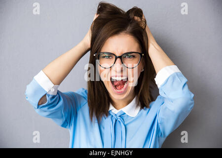 Angry businesswoman touching her hair and screaming over gray background. Wearing in blue shirt and glasses. Looking at camera Stock Photo