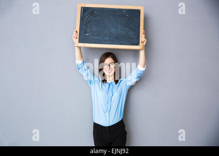 Happy woman standing with billboard over gray background. Wearing in blue shirt and glasses. Looking at camera Stock Photo