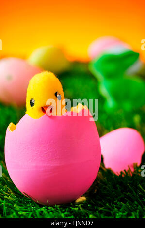 a teddy chick emerging from a pink easter egg on the grass and a green easter rabbit and a pile of colorful decorated eggs in th Stock Photo