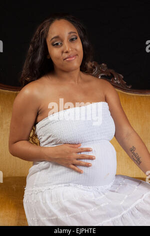 Pretty Black woman who is pregnant and wearing a white dress, relaxing on a gold couch Stock Photo