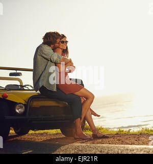 Young couple in love embracing and kissing. Young man and woman sitting on their car hood. Romantic young couple on road trip. Stock Photo