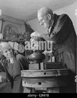 Sculptor William Snell seen here at work in his studio being watch by his schoolgirl model as he adds the finishing touch to a carving. 16th October 1952 Stock Photo