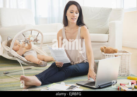 Mixed race mother paying bills on laptop with baby on floor Stock Photo
