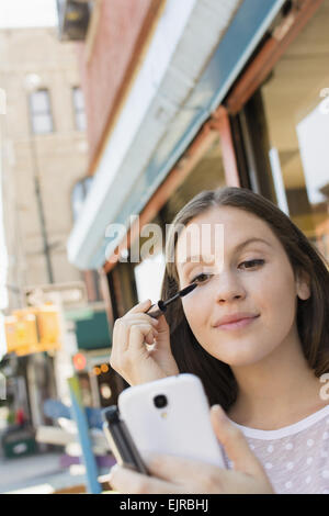 Caucasian woman applying makeup in cell phone camera Stock Photo