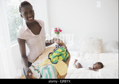 Black mother carrying laundry near sleeping baby boy in bedroom Stock Photo
