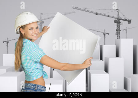 Girl in helmet showing empty paper sheet. White cubes with tower cranes Stock Photo