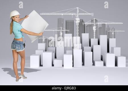 Girl in helmet showing empty paper sheet. Full lenght. White cubes with tower cranes Stock Photo