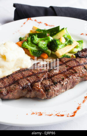 A white plate holds a delicious grilled New York Strip Steak with a side of mashed potatoes and fresh mixed vegetables. Stock Photo