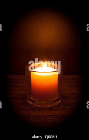 A candle glows in the darkness on top of sheet music; dark vignette Stock Photo