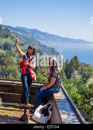 A young female takes a selfie with her smartphone with the coastline of the Pacific Ocean and the mountains at Big Sur, CA. Stock Photo