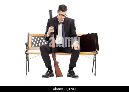 Depressed young businessman holding a shotgun rifle and looking down seated on a wooden bench isolated on white background Stock Photo