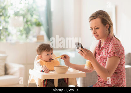 Mother feeding little boy (2-3 years) in high chair and texting message Stock Photo