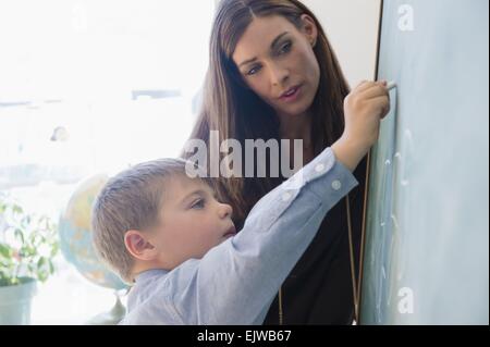 Female teacher assisting schoolboy (6-7) while writing on blackboard Stock Photo