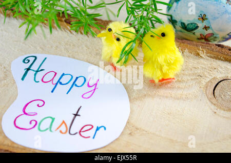 Two little yellow chickens standing beside a Happy Easter card on a wooden table with green plants in the background Stock Photo