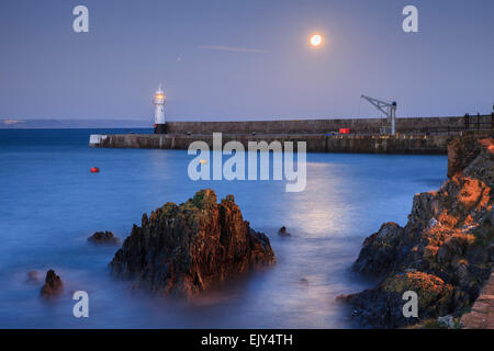 The Moon reflected in the outer harbour at Mevagissey in Cornwall, with the pier and lighthouse in the distance, Stock Photo