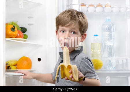 Little cute boy eating banana near open fridge Stock Photo