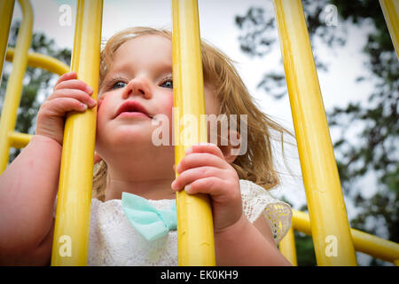 A small, blond toddler peeking through the bars of a jungle gym at a park playground. Stock Photo