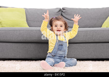 Playful baby girl sitting on the floor next to a modern sofa and gesturing happiness isolated on white background Stock Photo