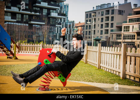 Young man reliving his childhood plying in a children's playground riding on a colorful red spring seat with a happy smile in an Stock Photo