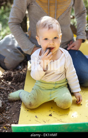 Little boy biting a cone while sitting on a touristic mat on the ground, mother behind the boy Stock Photo