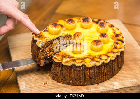 Slice being removed on a sharp kitchen knife and served from a freshly cut traditional home baked Easter simnel cake on a wooden chopping board Stock Photo