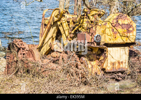 Abandoned and weathered yellow bulldozer crawler. Nature is trying to cover it with vegetation. Sea in background. Lots of rust Stock Photo
