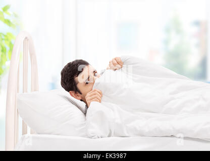 Scared guy lying in bed and hiding under a blanket at home Stock Photo