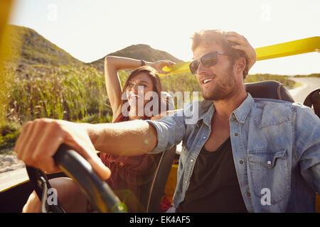 Cheerful young couple on road trip. Young man driving open topped car with woman smiling.