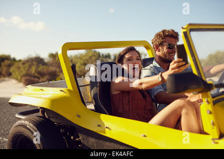 Excited happy couple enjoying road trip in their beach buggy. Man driving the car young woman taking a selfie from her mobile ph