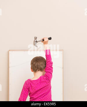 Little girl (5 years) holding hammer over her head and looking at artwork leaned against a wall. She is preparing to help with