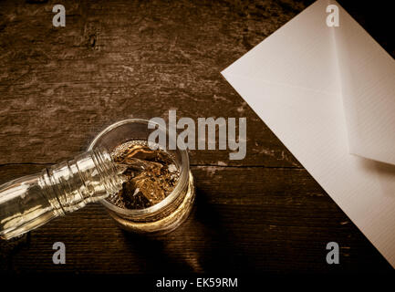 Close up of bottle pouring whiskey in a glass. The glass is placed on a dark rustic wooden table with a white envelope next to Stock Photo