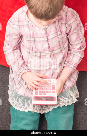 Little girl sitting on sofa at home with calculator in her hand. She is learning mathematics. Stock Photo