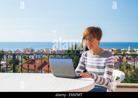 Mature lady with glasses and casual clothings working at laptop outdoors on terrace. Background of green hills and blue sky. Stock Photo