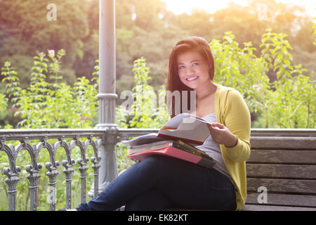 Beautiful young female university student with books and files on lap, sitting on wooden bench under pergola in a park. Stock Photo