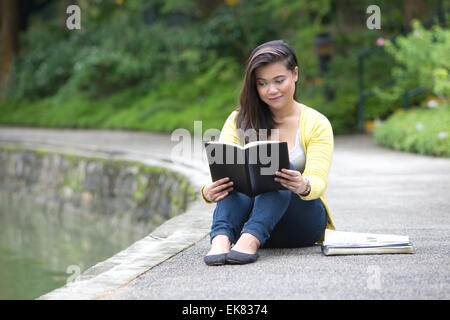Beautiful young female university or college student reading a book, seated by a lake in a park. Stock Photo