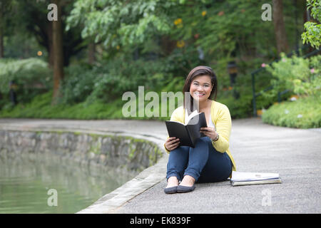 Beautiful young female university or college student smiling while holding a book, seated by a lake in a park. Stock Photo