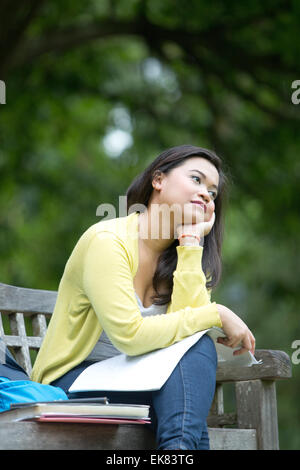 Young asian female student sitting on wooden seat in park,  books on lap and appearing thoughtful or pensive. Stock Photo