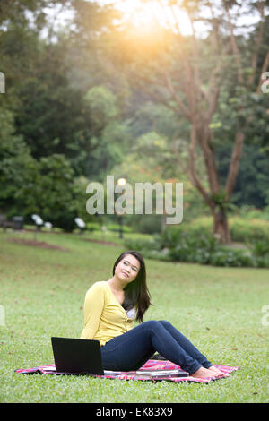 Beautiful young female university or college student thoughtful, with books and laptop, sitting in a park. Stock Photo